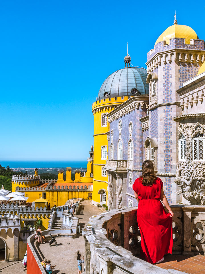 Girl in red dress on balcony overlooking Pena Palace and ocean.