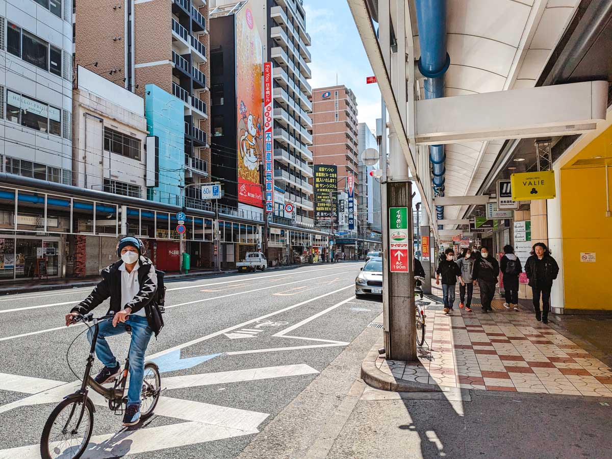 Man cycling down street of Den Den town next to covered sidewalk.