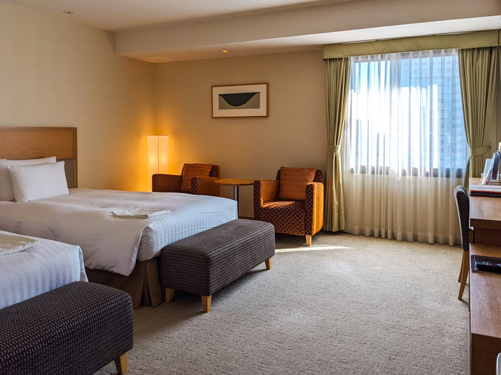 Interior of hotel room with two beds, desk, two chairs, and window.