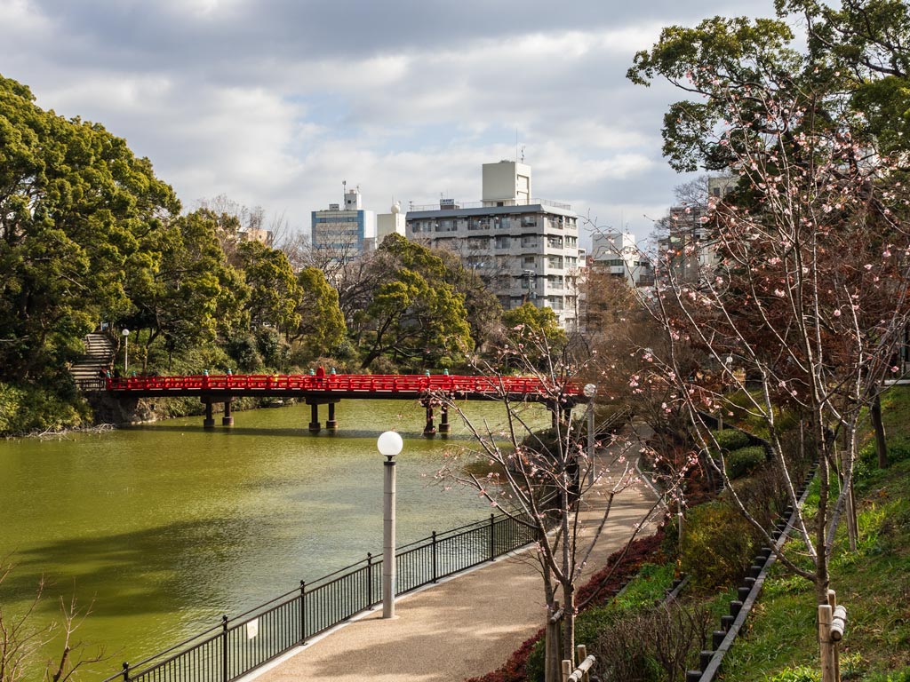 Osaka Tennoji Park view of pathway along river with red zen bridge crossing waterway.