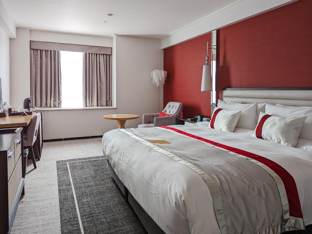 Inside of hotel room with large red and white bed, grey carpet, desk, dresser, and window.