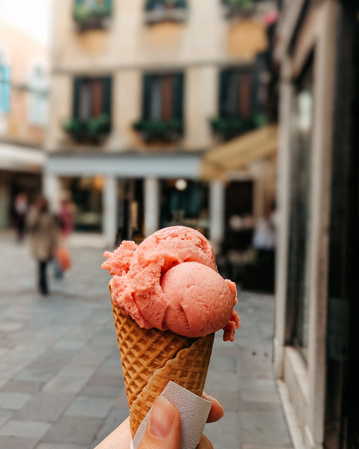 Strawberry gelato cone held in front of Venetian storefront