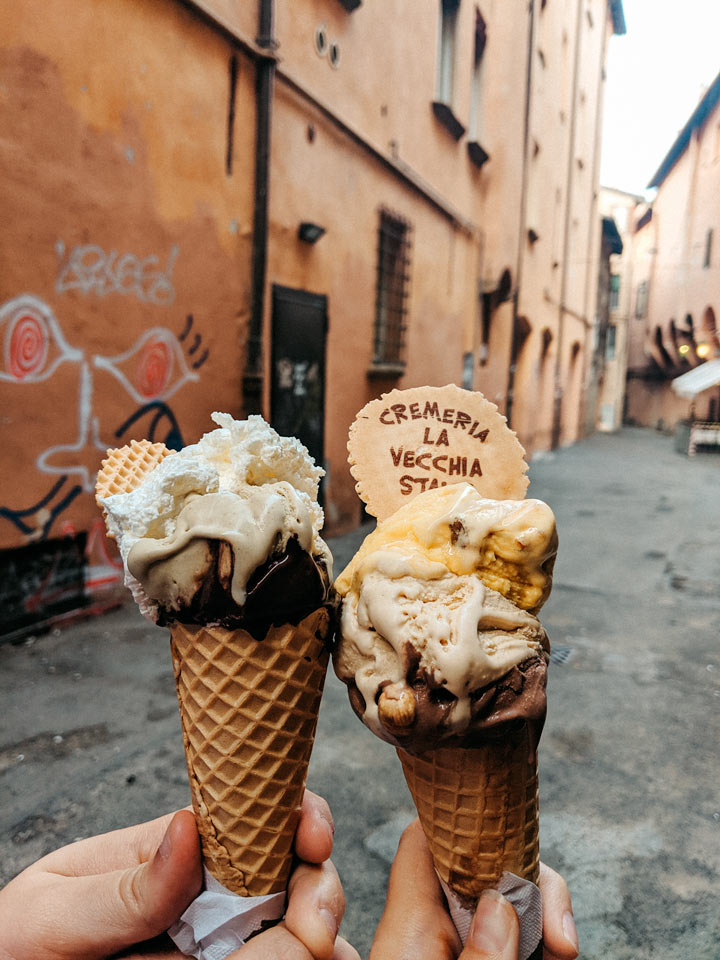 Two hands holding gelato cones with alley in the background.