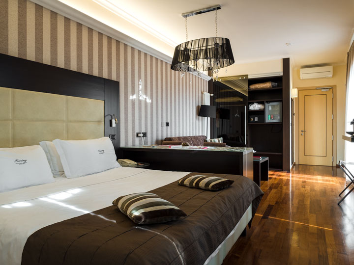 Interior of hotel room with large bed, hardwood floors, and black lamp hanging from ceiling.