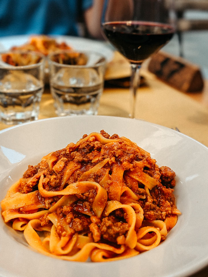 Plate of tagliatelle al ragu with red wine glass in background.