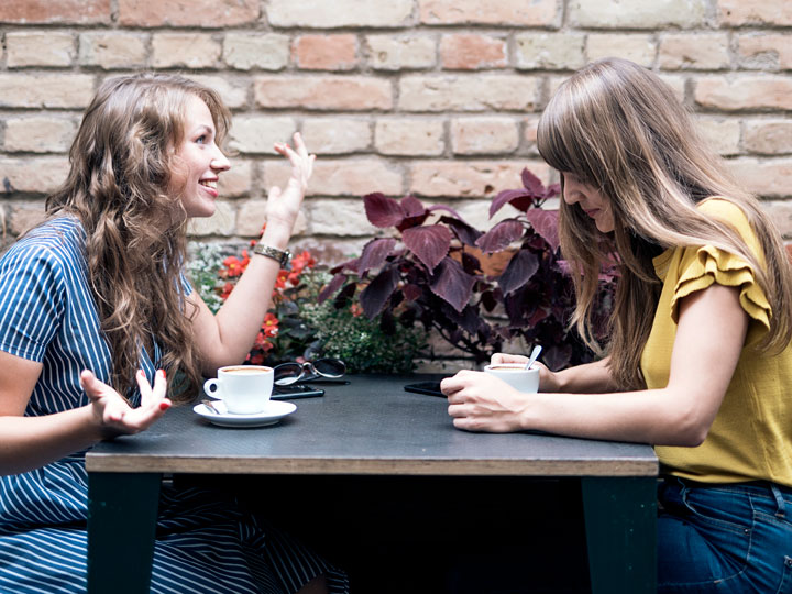 Two young women drinking coffee  at table and discussing advantages of living abroad.