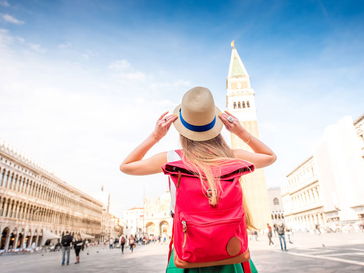 Girl with red backpack holding straw hat in Venice St. Mark's Square.