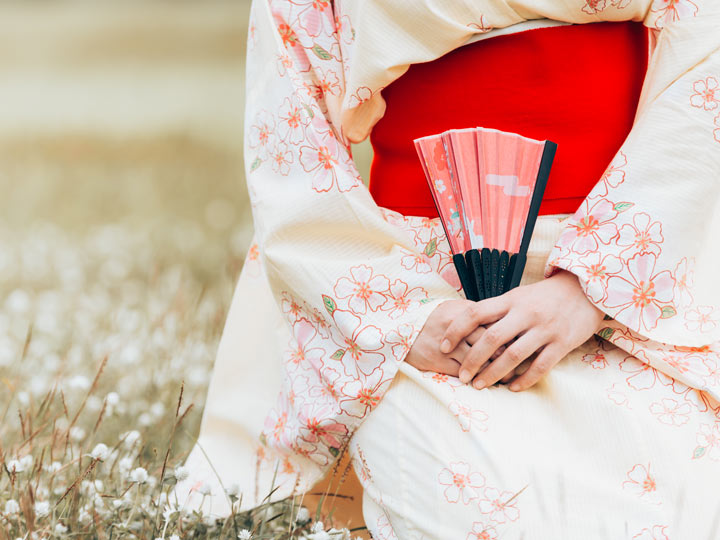 Woman in cream colored kimono sitting in field holding pink hand fan.