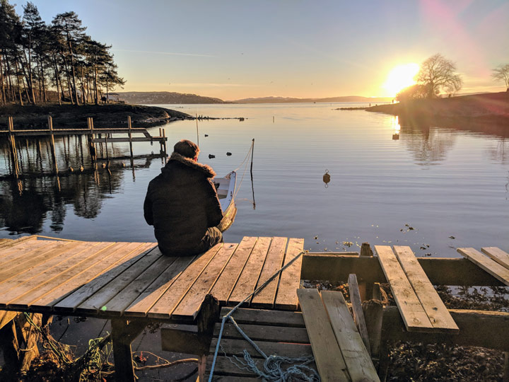 One person sitting on wooden pier overlooking water at sunrise during winter in Oslo Norway.