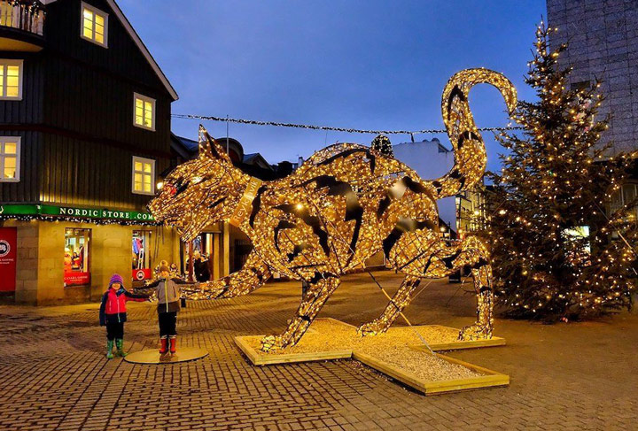 Two children standing near illuminated cat sculpture at night in Reykjavik during winter.