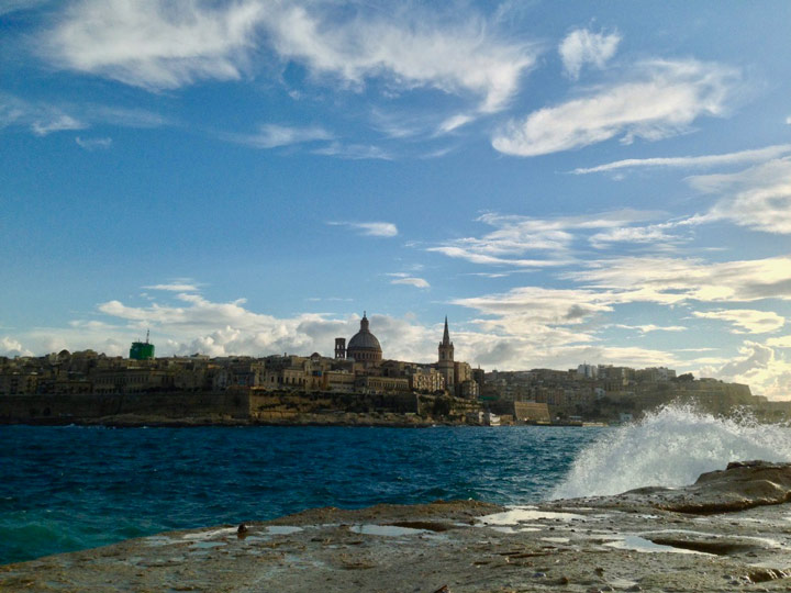 View of Valletta across water with crashing waves on sunny day.