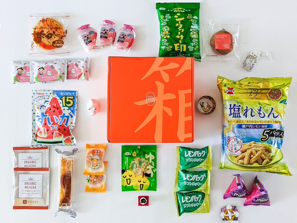 Flat lay of various Japanese snack packages on white table, with orange 2021 Bokksu review box in center.