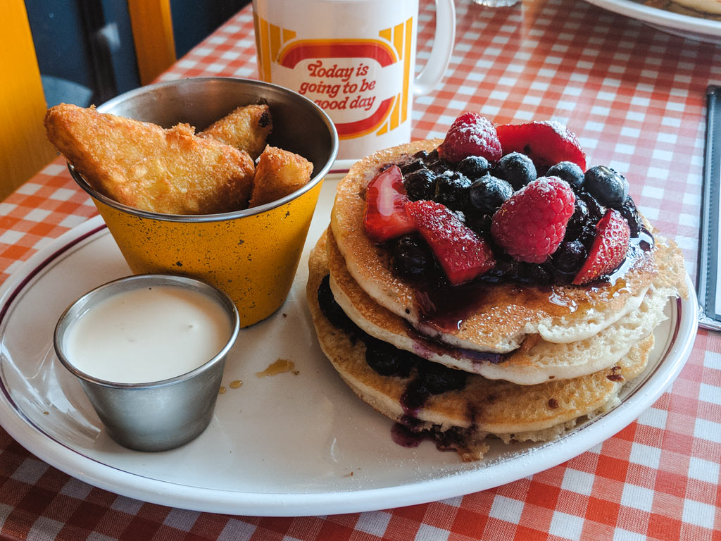 Plate of berry pancakes, hash browns, and cream on plaid tablecloth.