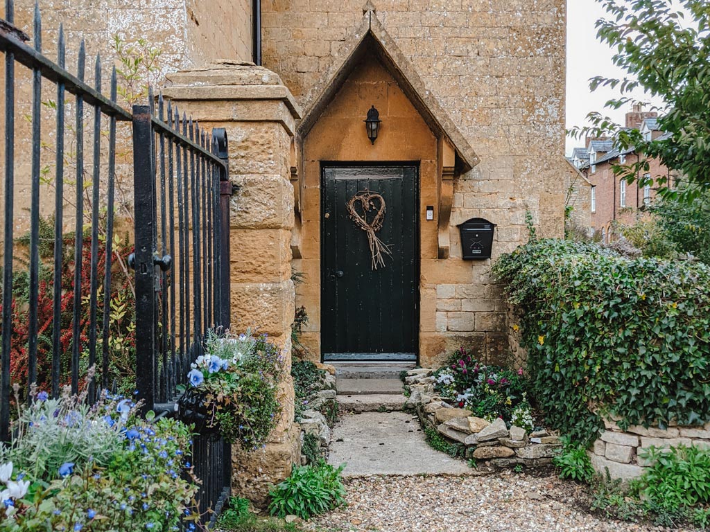 Yellow stone English cottage with black door and shrubs in front.