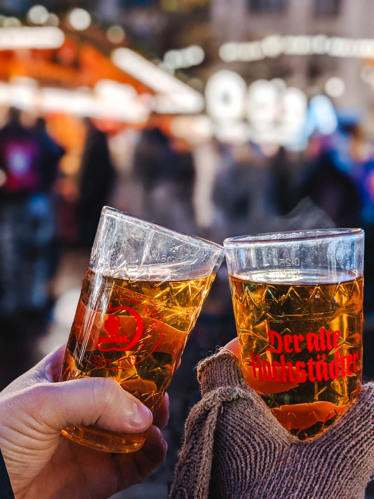 Two hands holding glasses of apfelwein with Frankfurt Christmas market in background.