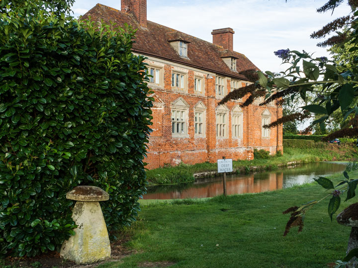 Old UK brick estate house with pond and green bushes in foreground.