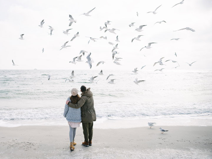 Couple on beach watching flock of seagulls fly overhead.