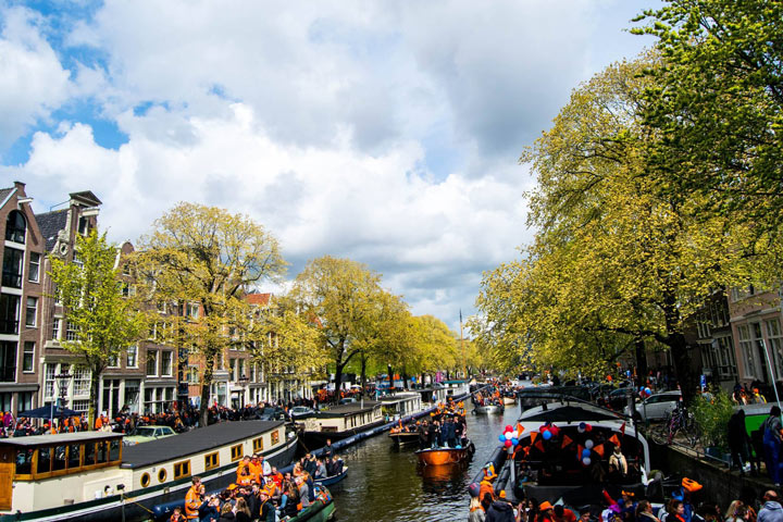 Amsterdam canal with tourist boats and lined with trees on partly cloudy day.