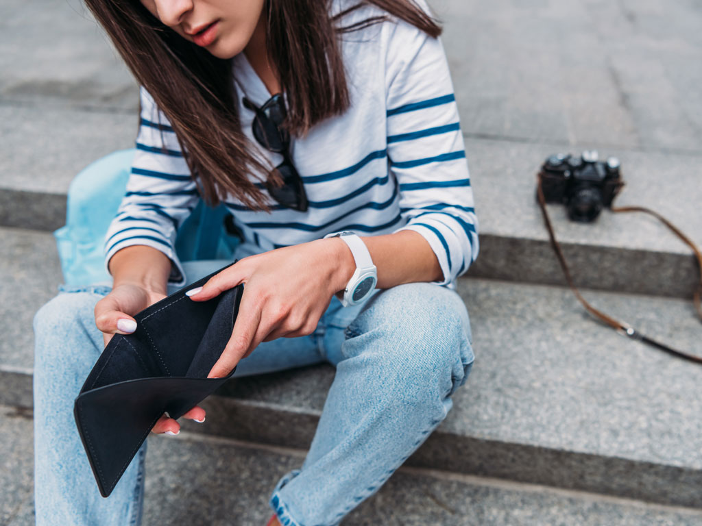 Young woman sitting on concrete steps, looking inside empty wallet after failing to save for a vacation.