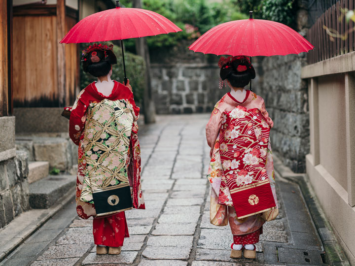 A geisha and maiko walking down street with red umbrellas.