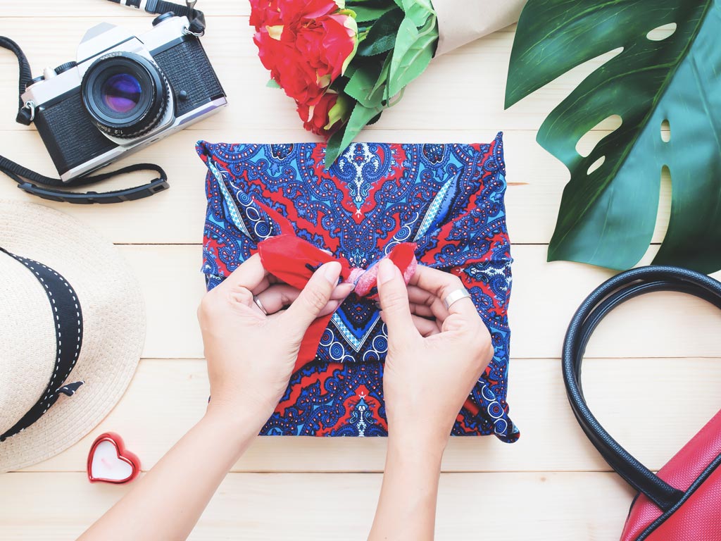 Woman wrapping present for someone moving abroad in paisley fabric, with camera, hat, and leaves on table.