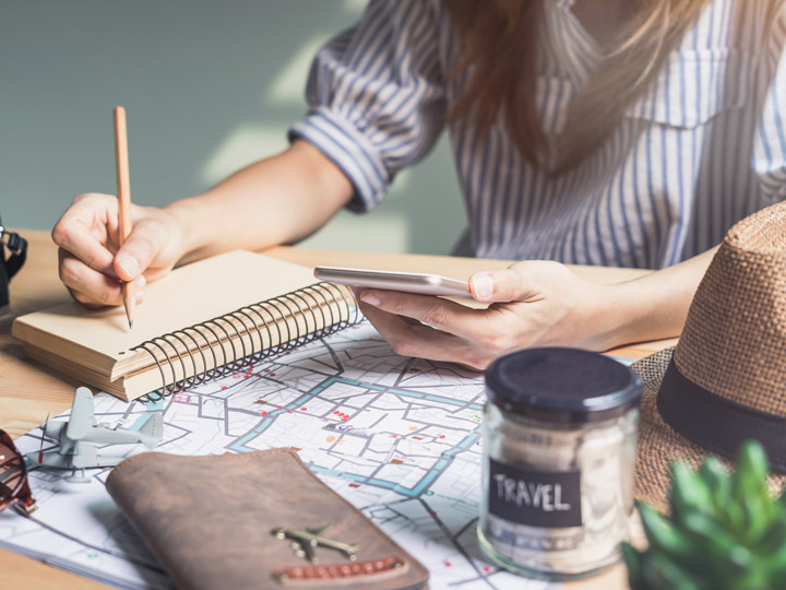 Girl sitting at desk holding phone and writing in notebook, with travel savings jar, map, and journal on desk.