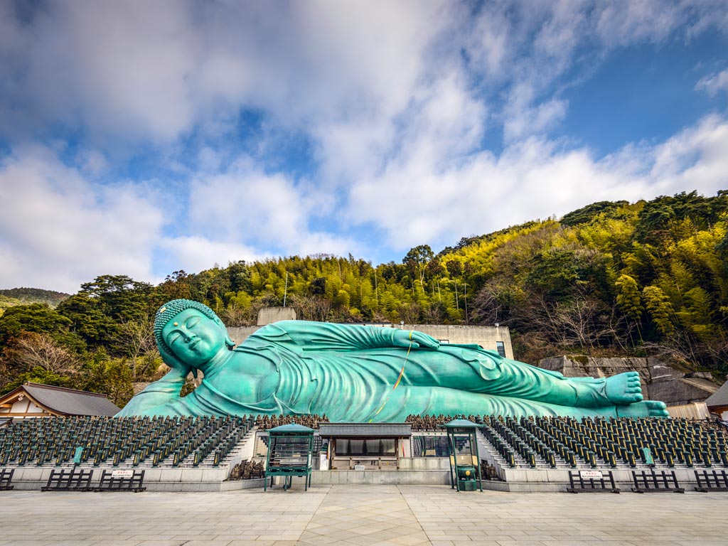 Large blue reclining Buddha statue with forested mountain in background.