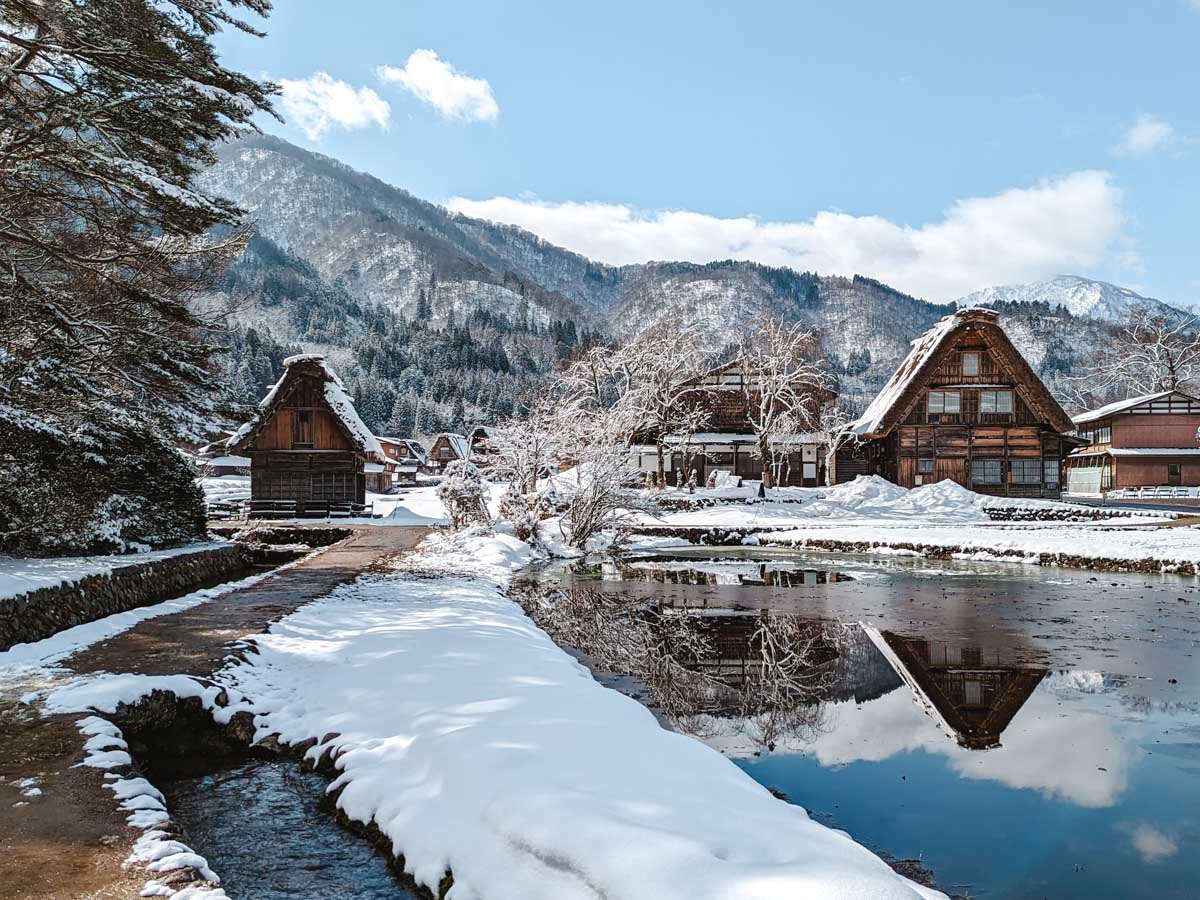 Shirakawago village traditional Gassho houses and reflecting pond amid snowy field and mountains.