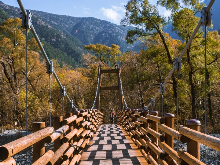 Man walking across wooden Myojin Bridge while hiking Kamikochi park in autumn.