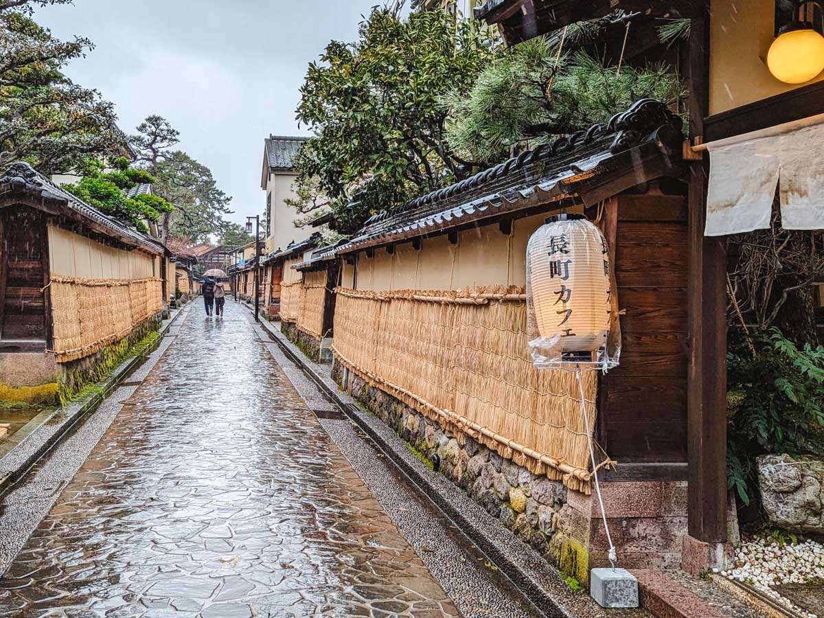 View of stone alley in Kanazawa Nagamachi with long walls and hanging lanterns in rain.