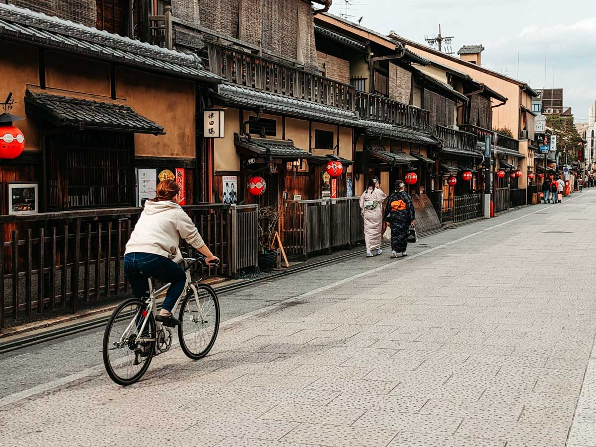 Woman cycling down Kyoto Gion alley lined with traditional Japanese buildings.