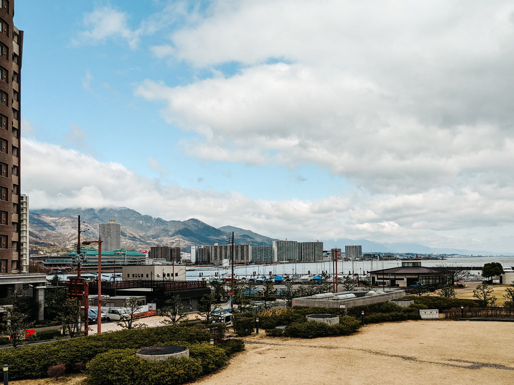 View of Otsu Port with ships and brown grass in foreground, and lake and mountains in the distance.