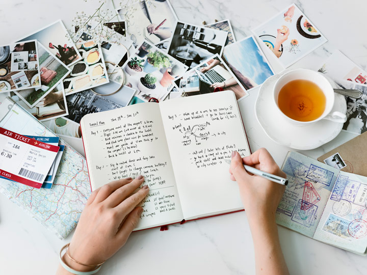 Person writing living abroad quotes in journal on white table with tea cup, photos, and plane tickets.