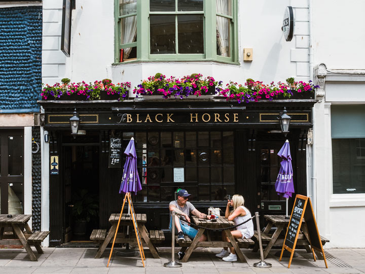 Two people discussing living in England pros and cons at table in front of Black Horse pub.