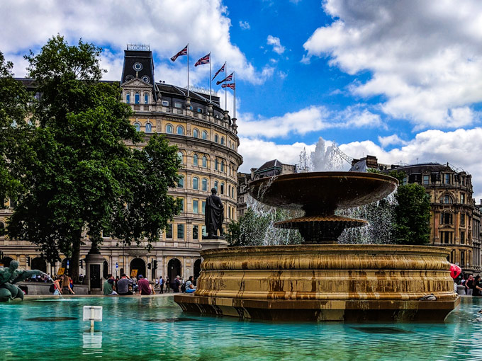 Fountain in Trafalgar Square with grand stone buildings in distance.