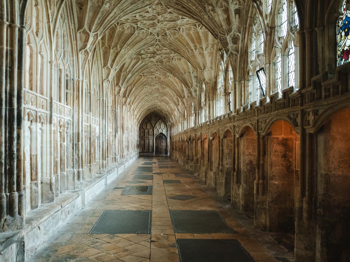 Interior corridor of Gloucester Cathedral with ornate ceiling details and stained glass windows.