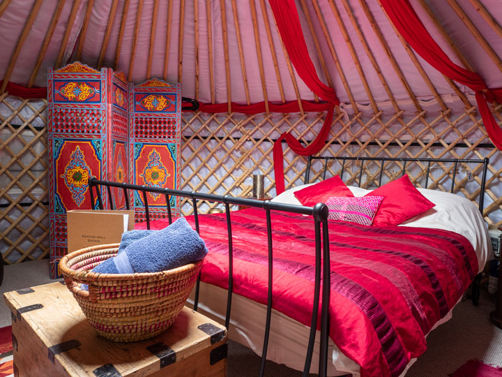 Yurt interior with red bed, wooden chest, towel basket, and screen.