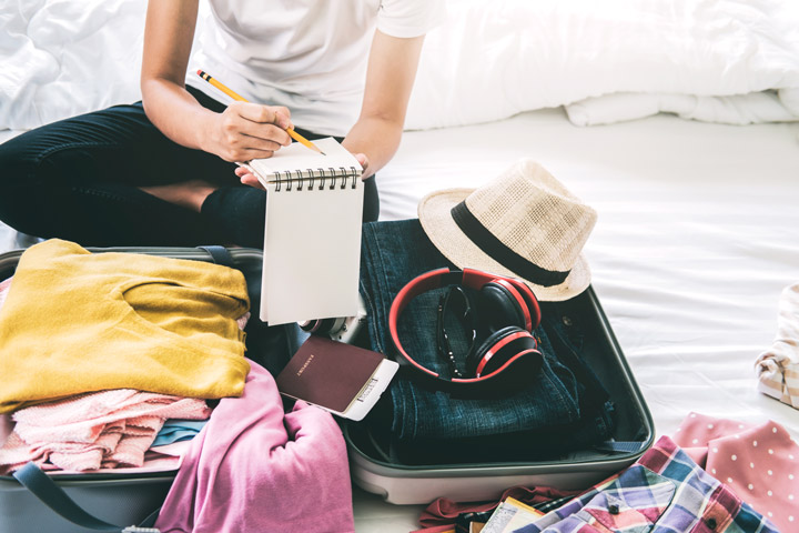 Girl writing moving abroad packing checklist on bed with full suitcase.