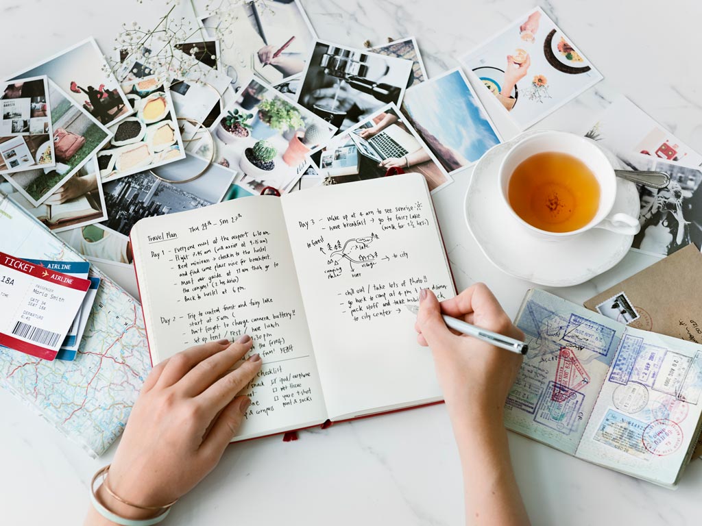 Hands writing in journal sitting on table next to tea cup, passport, and photos.