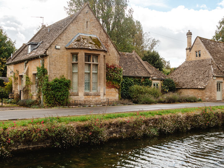 Stone cottage with ivy growing on front, behind flowing stream in Cotswolds