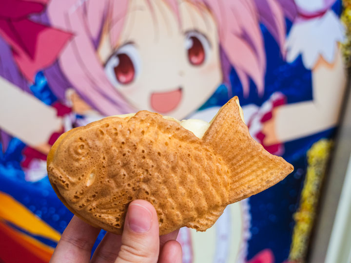 Fried fish pastry (taiyaki) held in front of anime mural.