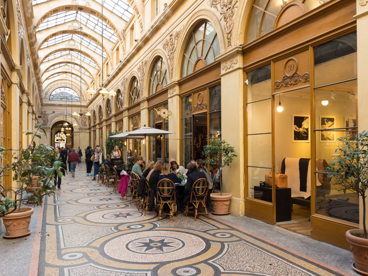 Inside of Galerie Vivienne covered passage with glass ceiling, mosaic floor, and cafe seating.