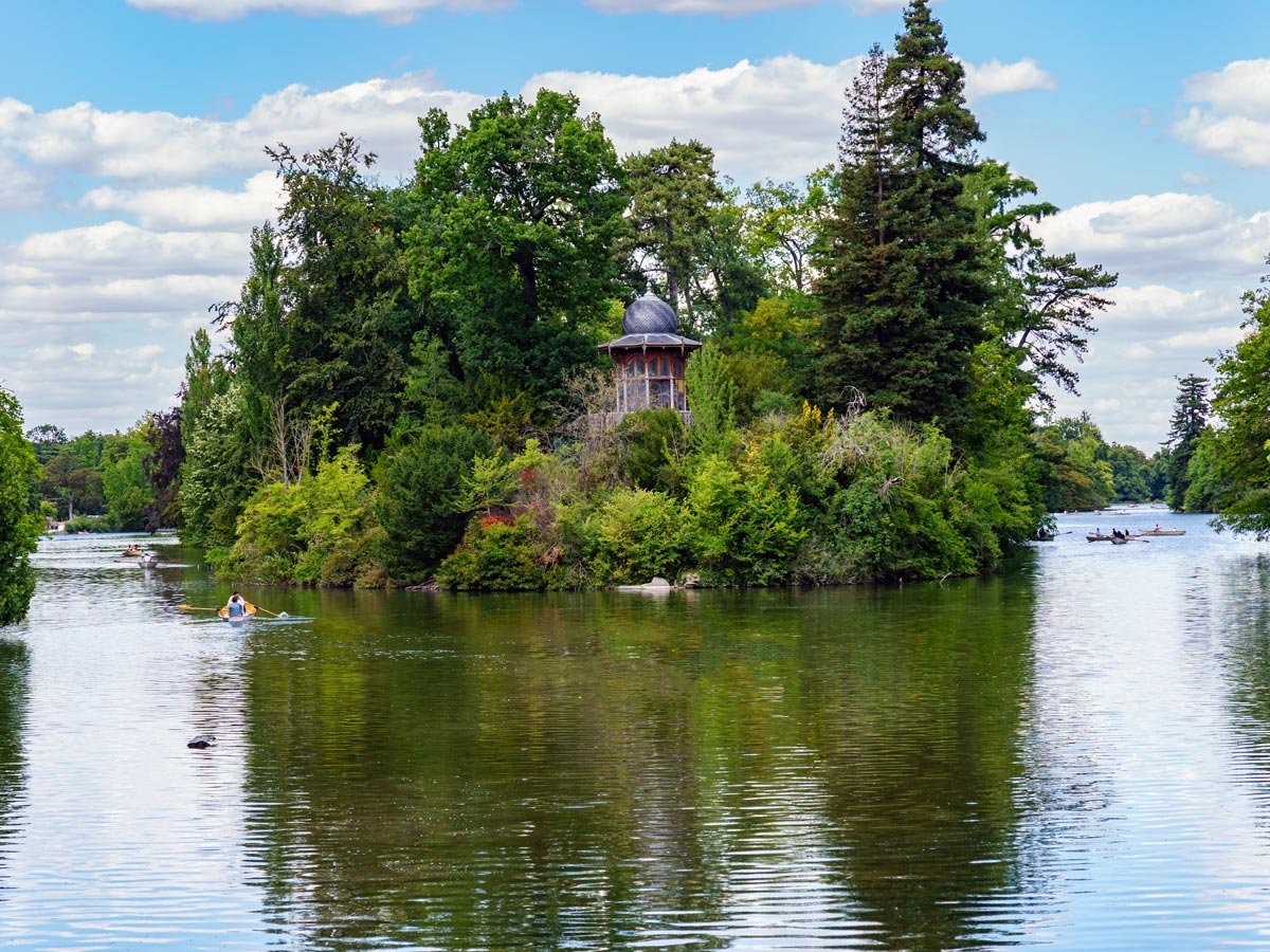 Bois de Boulogne park with pond of row boaters and small pavilion on green island.