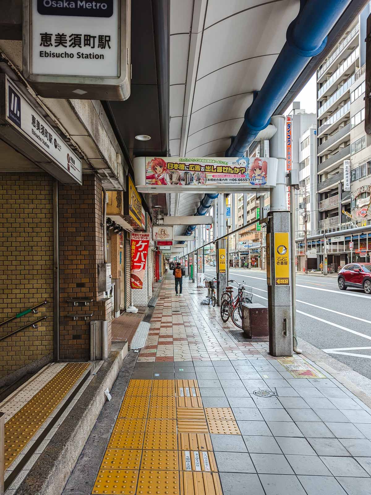 Osaka Den Den Town street outside Ebisucho Station exit with blue pipe along covered walkway.