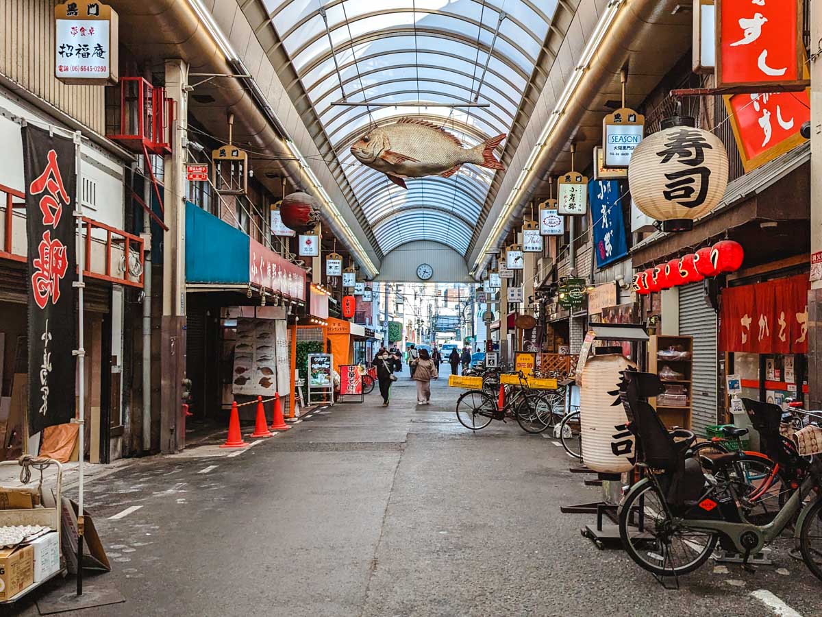 Inside covered shopping area of Kuromon Market with large white fish sculpture hanging from the ceiling.