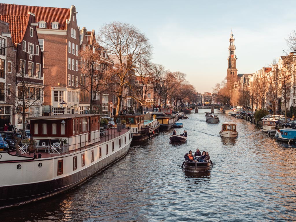 Small boats traveling down Amsterdam canal at sunset.
