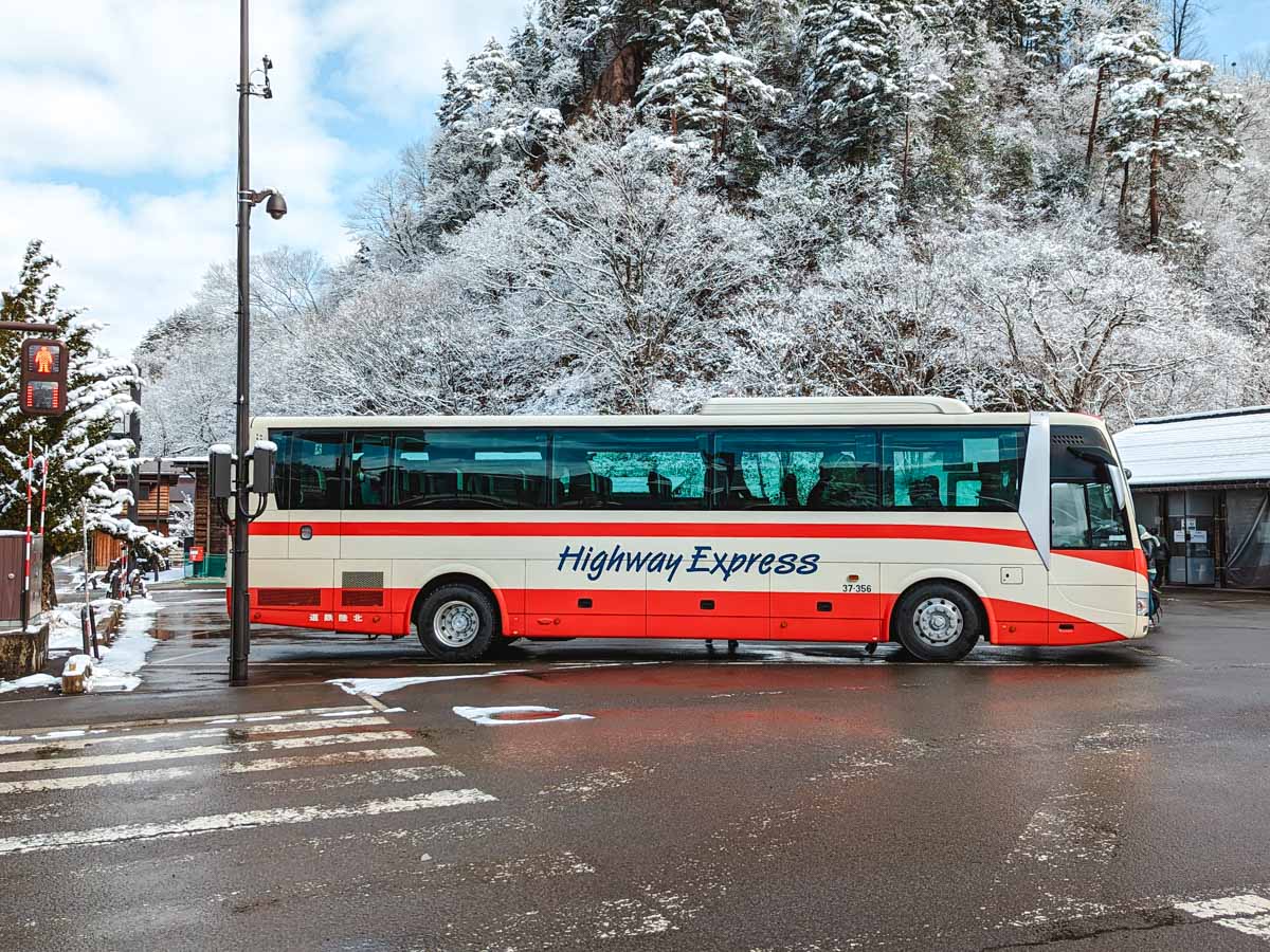 Red and white highway bus parked in Shirakawago bus terminal in front of snowy trees.