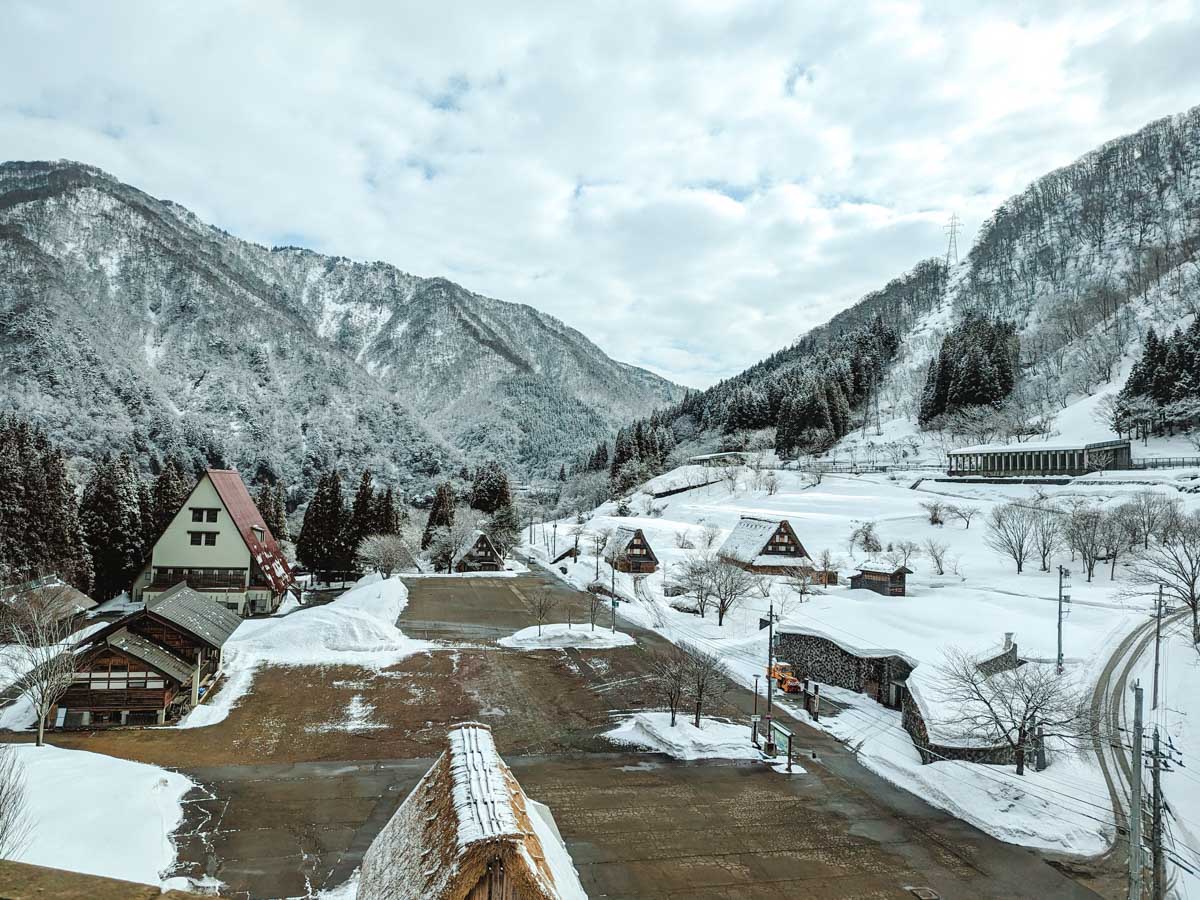 View of old Japanese houses and field covered in snow with mountains in distance.