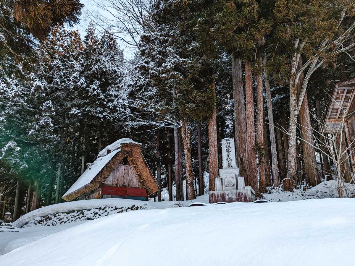 Small wooden outbuilding and grave marker amid yew trees and snow covered ground in Shirakawago village outskirts.