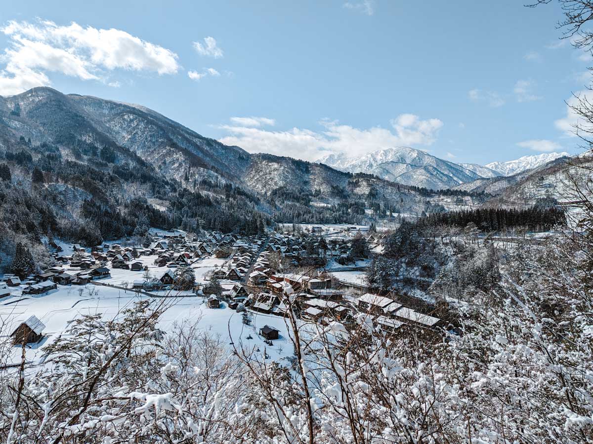 Panoramic view of Shirakawago village covered in snow with tree branches in foreground and mountains in distance.